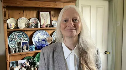 Linda Rogers at her home. She looks at the camera. Her long white her falls on her left shoulder and she has a grey blazer and white shirt on. Behind her is a Welsh dresser with a variety of ornaments displayed, including plates and framed pictures. 