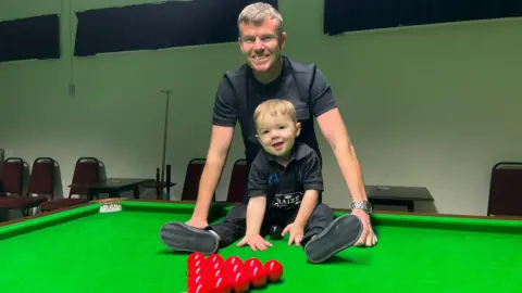 Jude is sitting on top of the snooker table with the balls in front him. His father Luke is standing behind him. They are both smiling at the camera. 