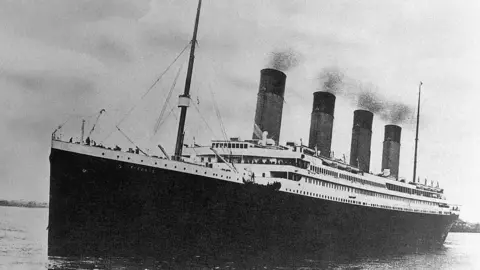 Getty Images A black and white photo of RMS Titanic embarking on its ill-fated maiden voyage. The large ship is dark in its lower part and white above deck, with four large funnels coming out the top.