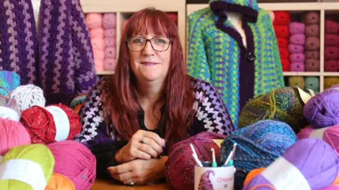 Rachael Mills in her home crochet studio- she is surrounded by colourful wool and crochet needles. She has long red hair and is wearing glasses.  