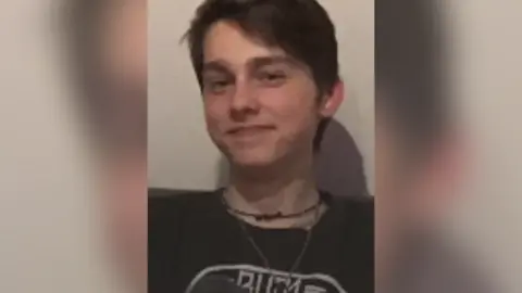 A teenage boy with brown hair styled with a fringe on the left covering part of his forehead looking into the camera. He is wearing a black t-shirt with a white logo, a small silver coloured chain and a black chain beneath it.