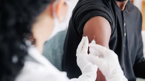 Getty Images A man wearing a black top rolls his sleeve up as he receives an injection in his upper arm. The woman administering the injection is in the foreground and is blurred. She is wearing a face mask, a white medical coat and white gloves. her hair is black.