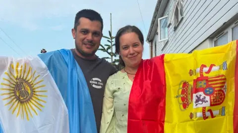 A man and a woman stand side by side holding their country's flags. The man has short dark hair and a beard and is wearing a grey top. He is holding the blue and white flag of Argentina, which is draped over his shoulder. Maria has shoulder-length hair and is wearing a yellow patterned shirt. She is holding the red and yellow flag of Spain.