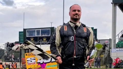 CWA A man with short hair and a beard, wearing a padded jacket, holding a black and white checkered flag and a trophy, stands in front of a stand of people on a race track.