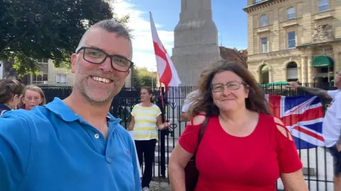 Ed Hill in a blue polo shirt and wearing black-framed glasses and Angie Nash wearing glasses and a red top. They are standing in front of the war memorial on Cathedral Green in Exeter with a Union flag visible behind them and a womanin the background holding a St George's Cross flag.