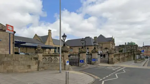 A Google streetview screenshot of Middlesbrough railway station. The large building has a red and white sign outside it, with gates for cars to access the car park. There is a forecourt to one side for pedestrian access.