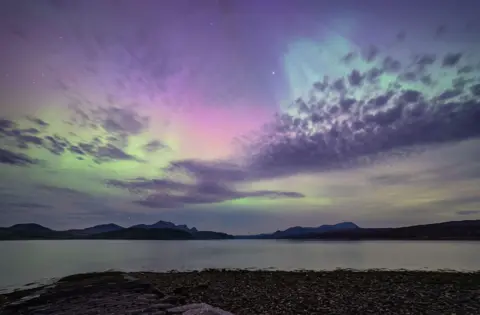 Gary Macleod The Northern Lights in the north coast of Scotland. There are green and purple stripes across the sky with mountains and the sea below. 