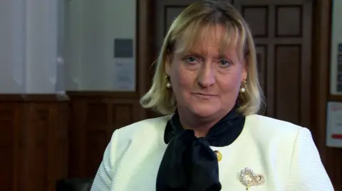 Nicola Connery is looking into the camera. She has short blond hair. She's wearing a white jacket with a pearl broach and a navy scarf. 