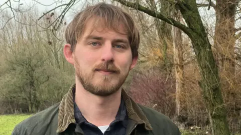 Alex looks at the camera with a small smile. He wears a green coat with a light brown collar and has light brown hair and a beard. 