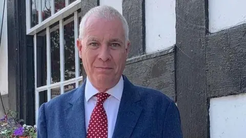Councillor Ewen Sinclair stands outside a building with a flower bed behind him. He has grey hair and is wearing a blue suit with a jacket and tie.