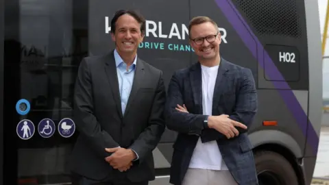 PA Media Mike Dawson (left), is standing smiling at the camera. he is wearing a black suit jacket and light blue shirt. He has brown hair. Gavin Jackson, right, is smiling at the camera and has his arms crossed. he is wearing a checked suit jacket and white t-shirt. He is wearing black framed glasses and has blondish hair. 