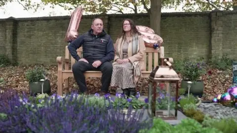 A man and a woman sit on a bench. They have two pink-silver balloons, shaped in a one and an eight. The man is wearing dark jeans and jumper with a black puffer gilet over the top. The woman is in a brown dress, boots, light brown coat and scarf. They are sat in front of a grave with purple flowers around it and a large lantern on it. Behind them there is a tall stone wall.