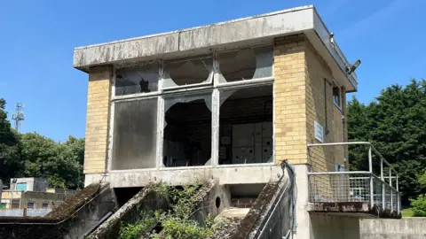 A small building housing machinery with yellow brick walls and three large windows, two of which have been smashed 