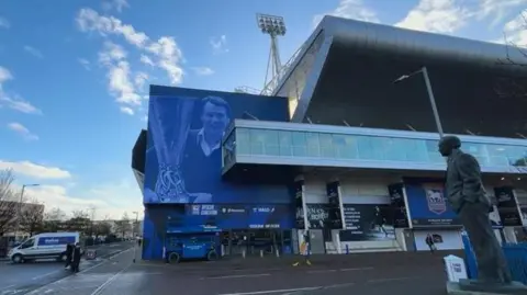 Luke Deal/BBC A wider view of Portman Road stadium with the new image of Sir Bobby Robson on the side of it. A statue can be seen to the right of the image. People walk near the stadium. A white van can be seen parked outside.