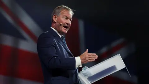 Reuters Rupert Lowe looks animated while standing at a lectern on stage, speaking through a hands-free microphone, with an image of a Union Jack behind him. 