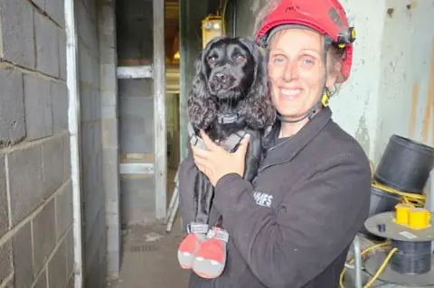 WMFS A woman in a red hard-hat and black jacket with the letters WMFS on it, holds up a black cocker spaniel who wears red and grey protection boots.