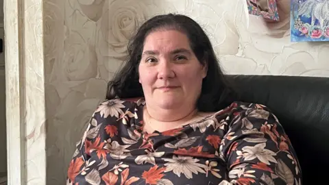 A woman in a floral dress sat on a leather chair and looking at the camera. She has a slight smile.