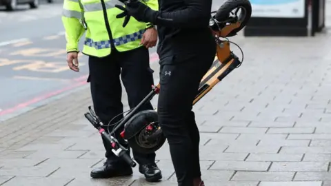 PA Media A man stands while holding a folded up e-scooter. A police officer stands beside him. Both are pictured from the neck down. 