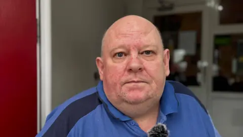 Justin Rogerson has short grey hair on the sides of his head and wears a blue sports t-shirt.