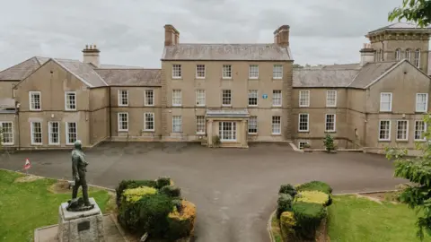 Royal School Dungannon. It is a large sandstone building with three stories in the main building and two stories the buildings connecting to it. The windows are large with white pain and lawns to the front with a statue on the left hand side.