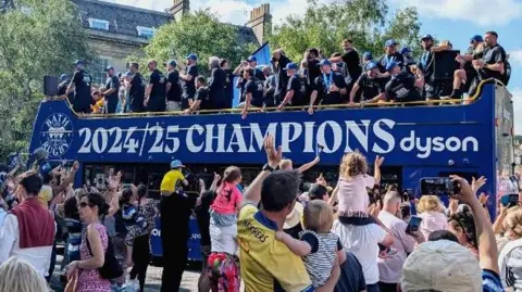 Blue bus with 2024/2025 champions written on the side. Bath players are on the top of the bus dressed in black t shirts with blue Bath caps. Lots of people are waving and taking photos in the foreground.