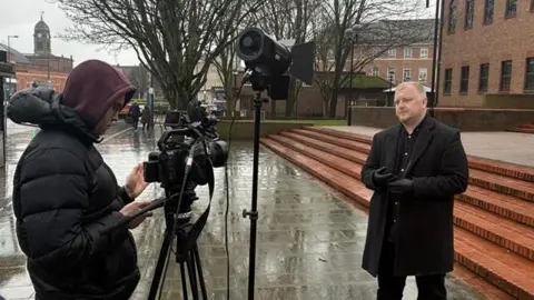 A man in dark clothing stood outside of Derby Crown Court presenting to a camera crew