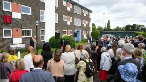 PA Media A view of a crowd of people taken from behind. They are watching a woman speaking from a podium outside a block of flats.