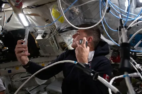 NASA This close interior image shows Canadian astronaut Jeremy Hansen floating inside the cramped Orion spacecraft while shaving. He wears a dark hooded top with a small Canadian flag patch on the sleeve and holds a razor up to his cheek. Around him, the cabin walls are lined with white padded panels, storage bags and equipment boxes. Blue and white cables loop across the frame, and bright strip lighting overhead makes the small space look crowded, practical and lived in.