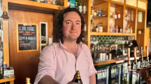 Peter Connolly is standing behind the bar at Nortons, holding a beer pump, wearing a pink and white striped shirt. 
