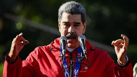 Getty Images President Nicolás Maduro in a red jacket, with a medal on. He has grey hair and a moustache. His arms are raised with fingers curled over, he is speaking into a microphone.