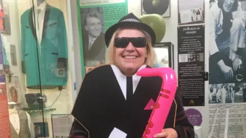 A woman with blond hair, a black hat and an inflatable pink saxophone standing behind a black and white board designed to look like a suit