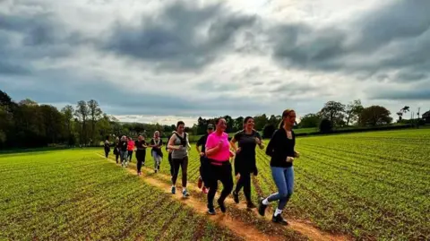 Runclub Woman run along a trail through farmland. There are trees in the background. Some of the runners are running side by side.