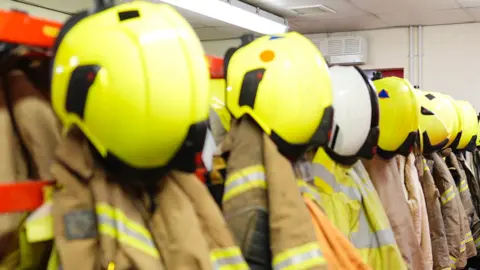 A picture of a number of jackets and helmets hung up in a row. Some are hi-vis and others are brown in colour.