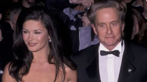 A dark haired woman in a shoulder-less dress and a man with wavy long hair in a tuxedo at a n event 
