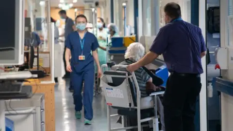 Patients and staff pictured in a hospital corridor, with an elderly women in a wheelchair in the foreground 