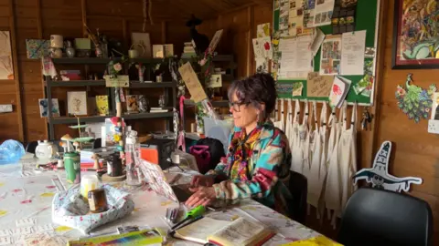 BBC/Helen Richardson Kylie is pictured inside her allotment shed. It is brightly coloured with pictures and images of mushrooms and fairies. She is typing on her laptop. 