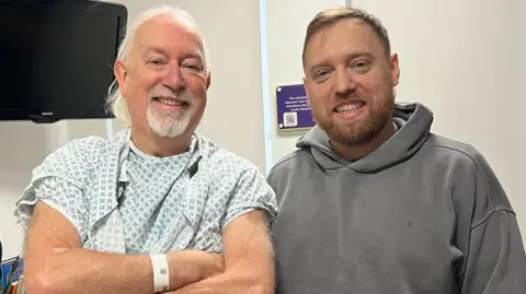 Suministrados dos hombres se paran uno al lado del otro mirando la cámara. Uno, con cabello blanco y barba, lleva un vestido de hospital y muñequeras que se le dan a los pacientes. El otro, con cabello castaño y barba, lleva una sudadera gris. En el fondo, hay un televisor en la pared y una planta en una superficie, pero la habitación está en su mayoría decorada en blanco.