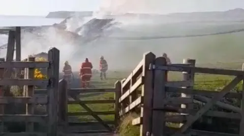 Three firefighters in red uniforms tackle a fire in the countryside behind a fence. Large plumes of smoke can be seen. 