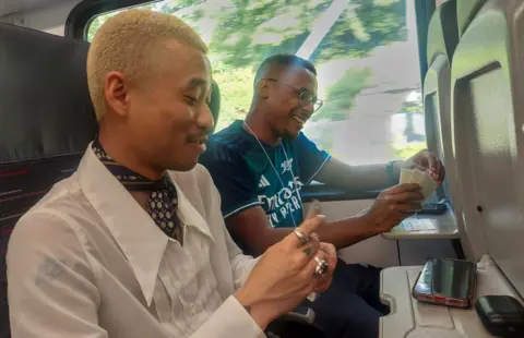 Two friends pass the time with a game of cards on board a train. One is wearing a white shirt and the other is wearing a dark blue Arsenal football shirt.