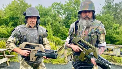 Two bearded men are wearing body armour, helmets and carrying replica guns all in khaki colours.
