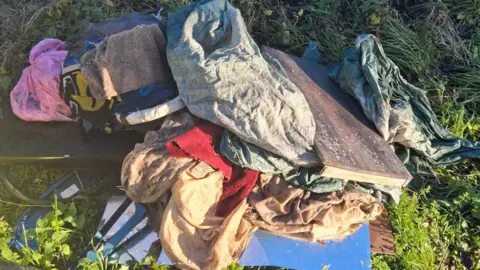 South Kesteven District Council Bin bags, fabric of various colours and a piece of brown wood dumped in a pile on a long patch of grass.