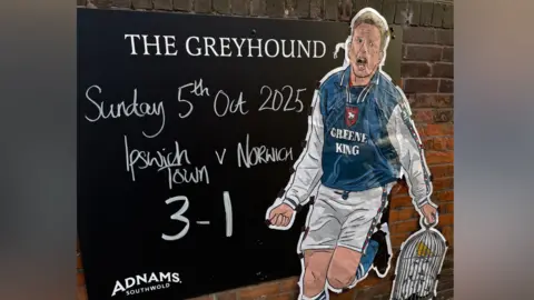 Dan Lightfoot/The Greyhound A blackboard on a red brick wall on the entrance to a pub. It has the pub's name, The Greyhound, painted on the top in white paint. Written in white chalk it then reads: Sunday 5th October 2025, Ipswich Town v Norwich, 3-1. There is a painting of a Town player celebrating as he holds a canary bird cage in it to represent Norwich City.