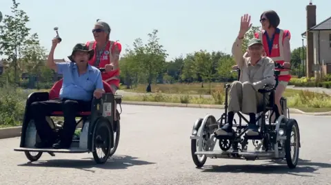 Two specially adapted trikes are being cycled, side-by-side, along a street. The trikes have two wheels at the front, wide enough apart to carry a passenger. They are powered and steered by women on the back, each wearing pink hi-viz vests. The passenger in the front of the trike on the left is an elderly man wearing a blue shirt and green baggy sun hat. He is holding a squeezy horn in the air.  The passenger in the trike on the right is an elderly man wearing a tan cap - he is sat in a wheelchair which is mounted on the trike. He is smiling and waving.