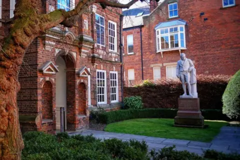 Hull City Council Wilberforce House is a brick building with 10 windows and an entrance. There are lawns, bushes, a tree and paving. There is a statue and a brick building in the background.