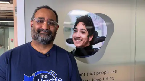 A man with a grey beard and glasses wearing a navy T-shirt is standing in front of a screen which has an image of a young man on it who is disabled.