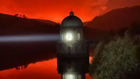 MWWFRS Rhayader A lighthouse is pictured in the water, shining a light. Above it is a red sky, lit up by wildfires in the distance. The red colour is also reflected in the water.