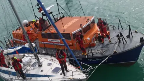 Jersey Lifeboat Association A large orange lifeboat pulled alongside a small white yacht. Crew members stand across both decks. The water is calm.