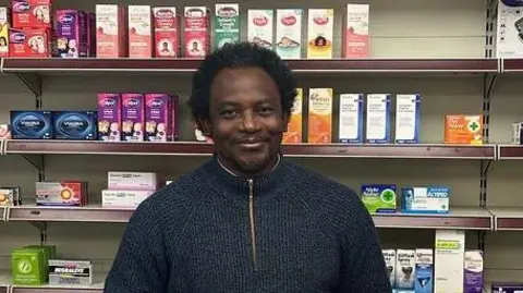 Tunde Olawoye Tunde Olawoye stood behind the counter at his pharmacy wearing a blue fleece jumper. There are shelves of drugs and medicine behind him.