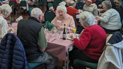 A group of elderly people sat around a table with glasses of wine and plates of food