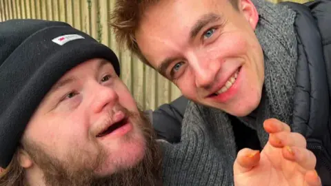 William Hessey Two men are in the image. One has Down's syndrome. He is wearing a black beanie hat and has a brown beard. The other man is smiling at the camera. He is wearing a coat and grey scarf. He has dark hair. 
